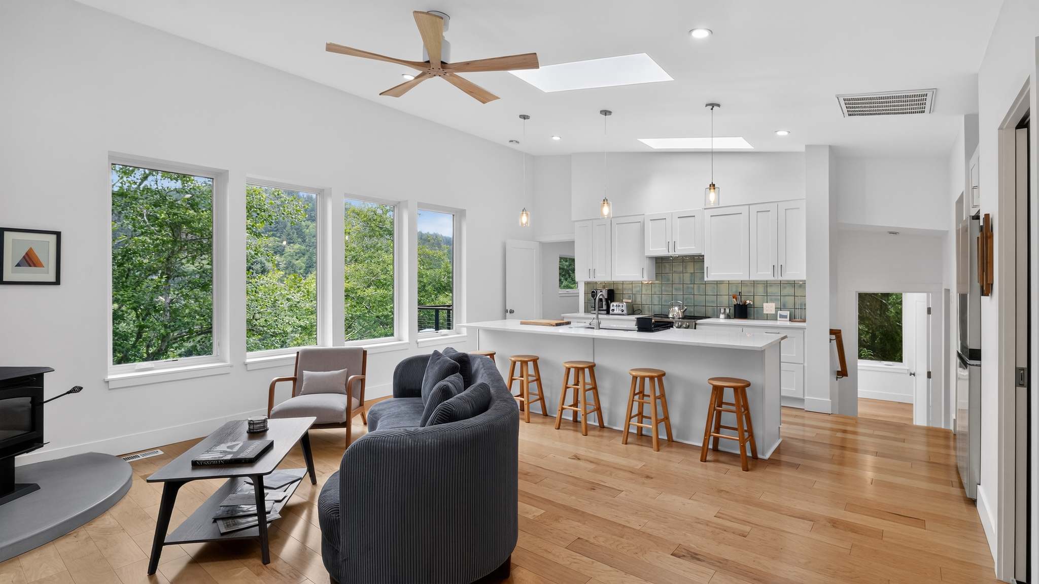 Wide shot of an open-concept living room and kitchen, featuring a gray curved sofa, a wood-burning stove, and a kitchen island with bar stools, all under high ceilings with skylights.