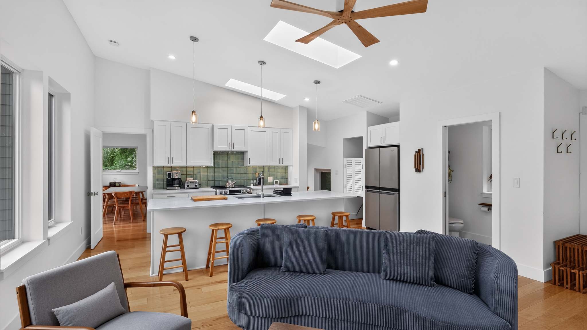 Modern kitchen with white shaker cabinets, a white island with a sink, and a green tiled backsplash, open to the living area with hardwood floors and a view of a refrigerator.