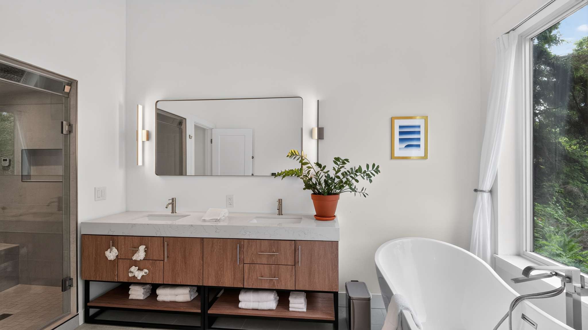 A bright, modern bathroom features a double vanity with a light wood cabinet, white quartz countertop, and two sinks. A large rectangular mirror with vertical LED lights hangs above, opposite a glass-enclosed shower and a freestanding white tub.