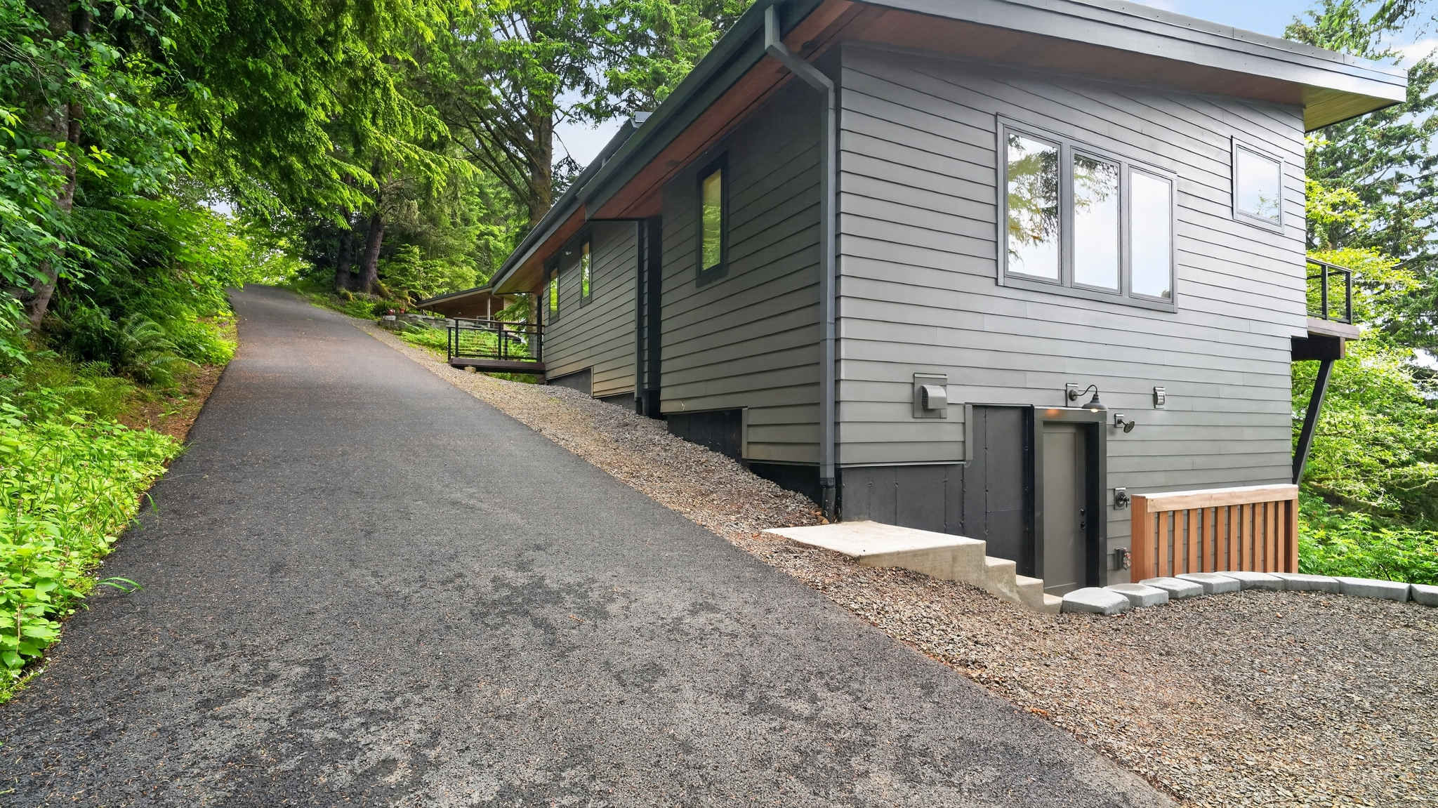 The side of a modern, dark gray house with horizontal siding and multiple windows is visible next to a steep asphalt driveway. A lower-level entrance with concrete steps and a wooden fence is nestled into the hillside.