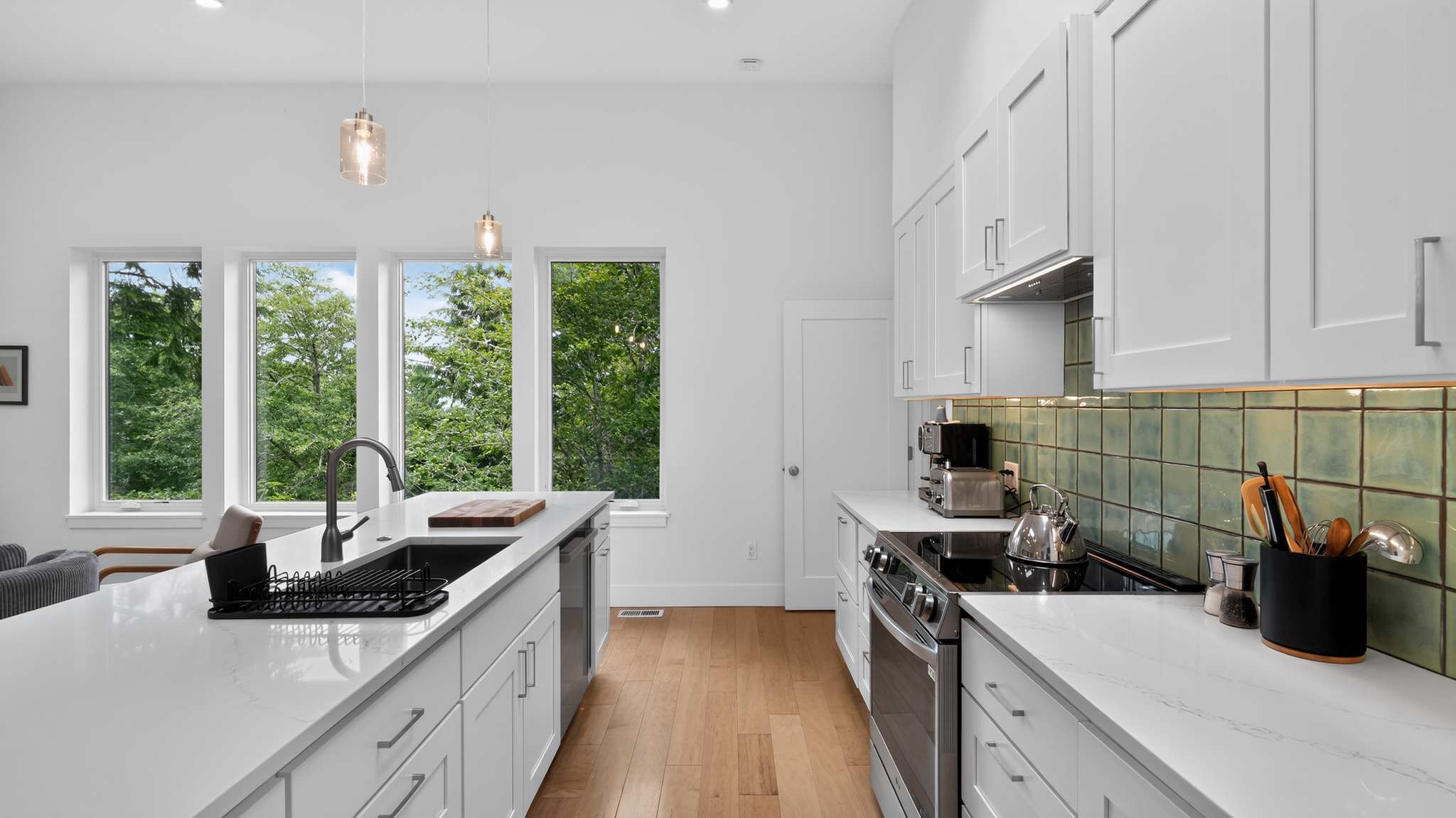 Bright kitchen with white cabinets, white quartz countertops, and a green tiled backsplash. A black sink is integrated into the island, and large windows offer views of lush trees. Stainless steel appliances are visible, including a dishwasher and range.
