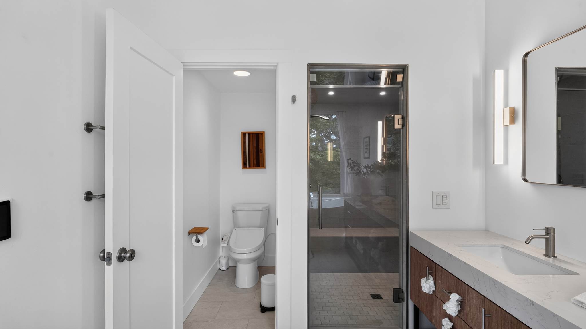 View into a modern bathroom with a glass-enclosed walk-in shower on the right and a separate toilet room with a bidet toilet on the left, both featuring light-colored walls and contemporary fixtures.
