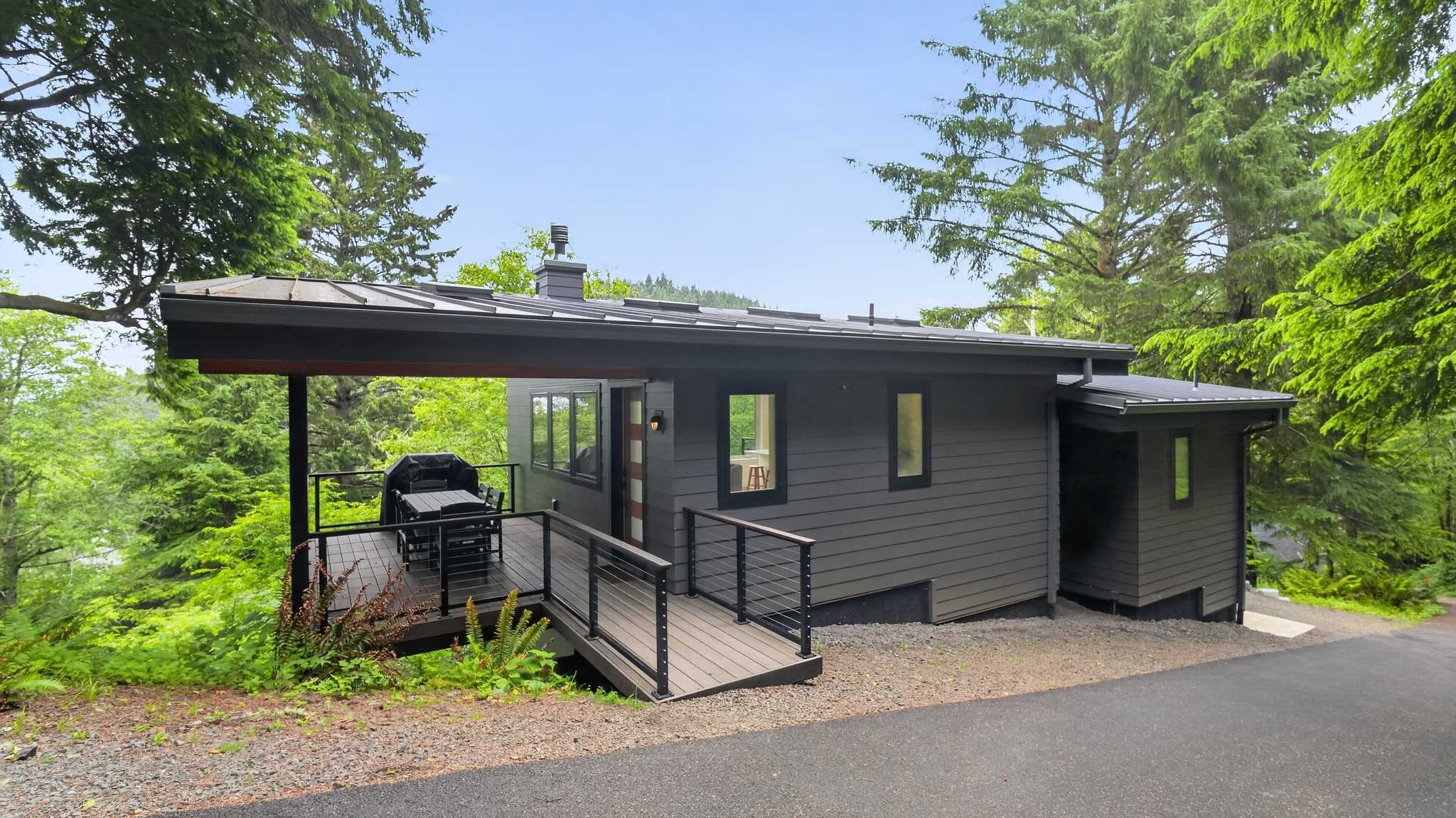 A modern, dark gray house with a sloped metal roof features a covered deck with a grill and a wooden ramp leading to the entrance. Tall evergreen trees surround the property, which is set on a hillside.