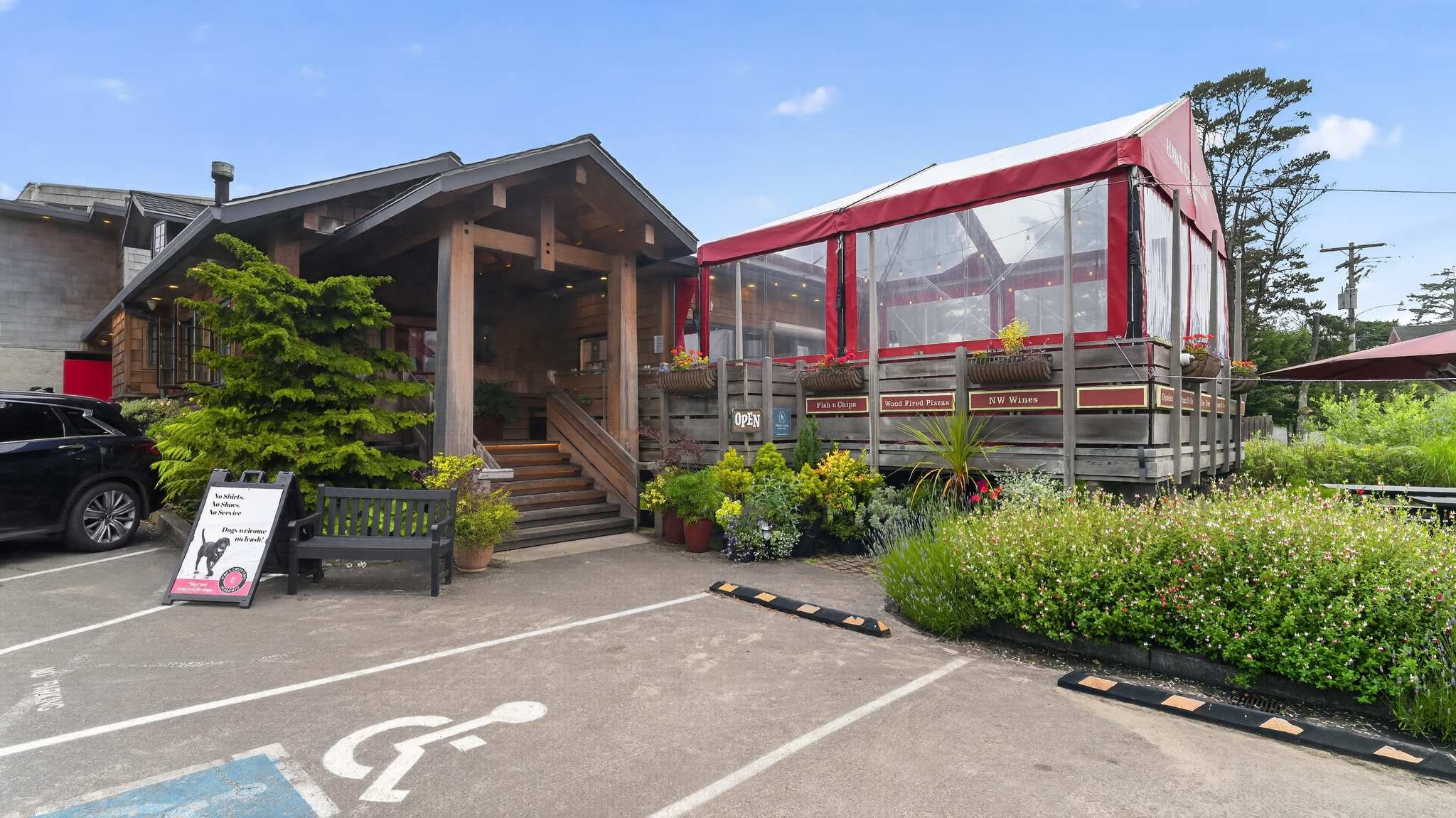 A rustic restaurant building with a wooden facade and a large outdoor dining area covered by a red and clear tent. Planters with flowers and greenery adorn the exterior, with a paved parking lot in the foreground.