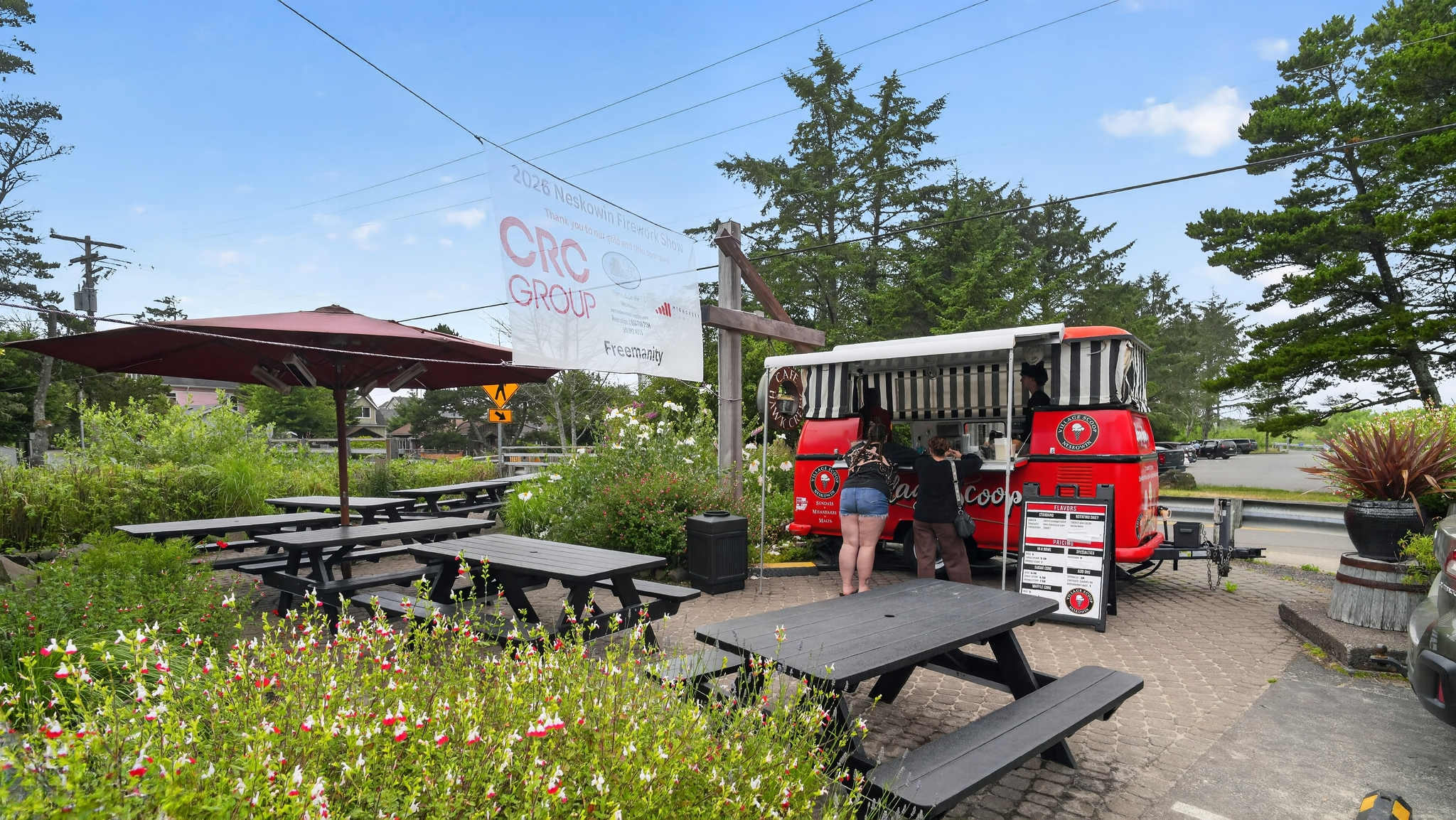 A vibrant red food truck with black and white striped awnings is parked next to several black picnic tables with umbrellas, surrounded by flowering bushes and trees under a clear blue sky. People are ordering from the truck.