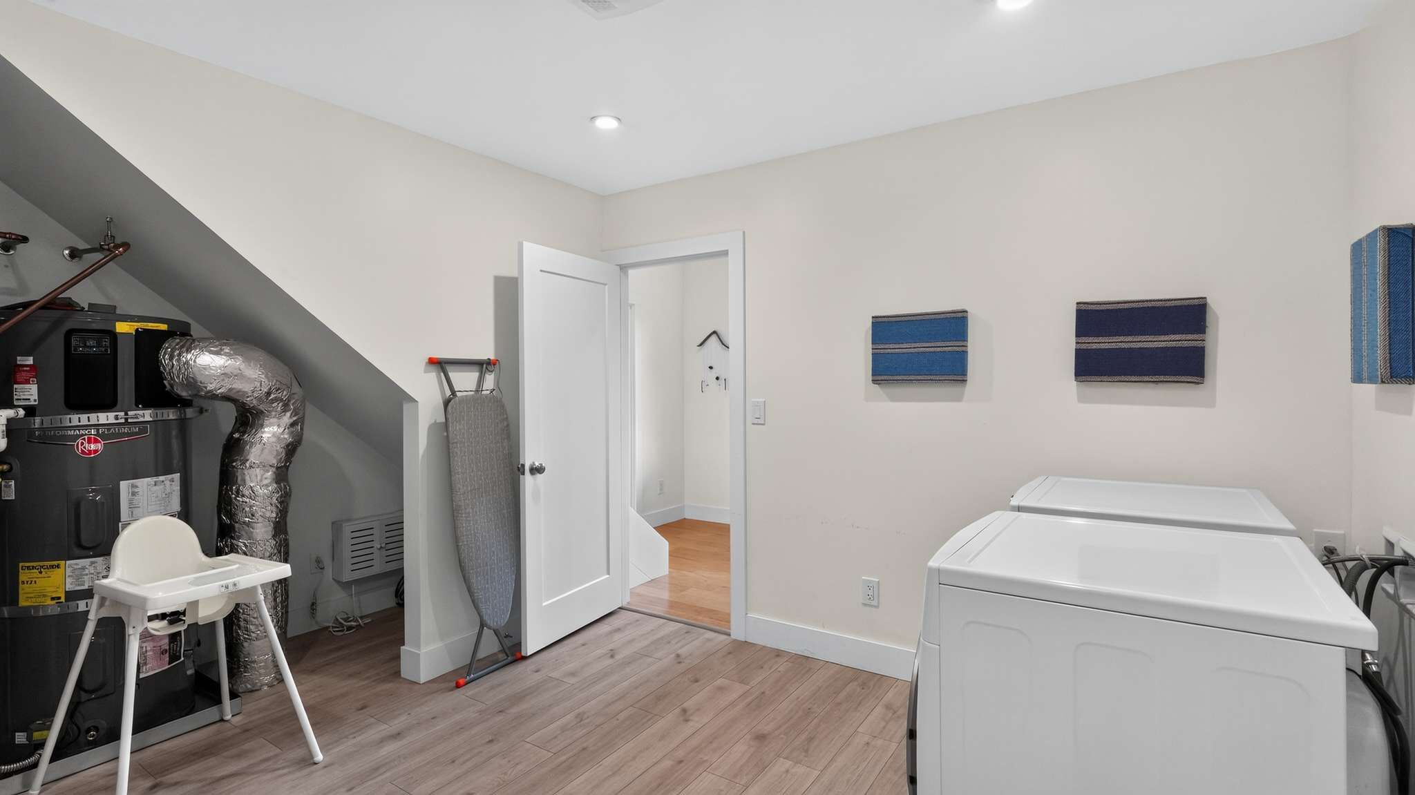 A utility room serves as a laundry area, featuring a water heater with silver insulation, a white front-loading washer and dryer, and an ironing board. The room has light wood-look flooring and white walls.