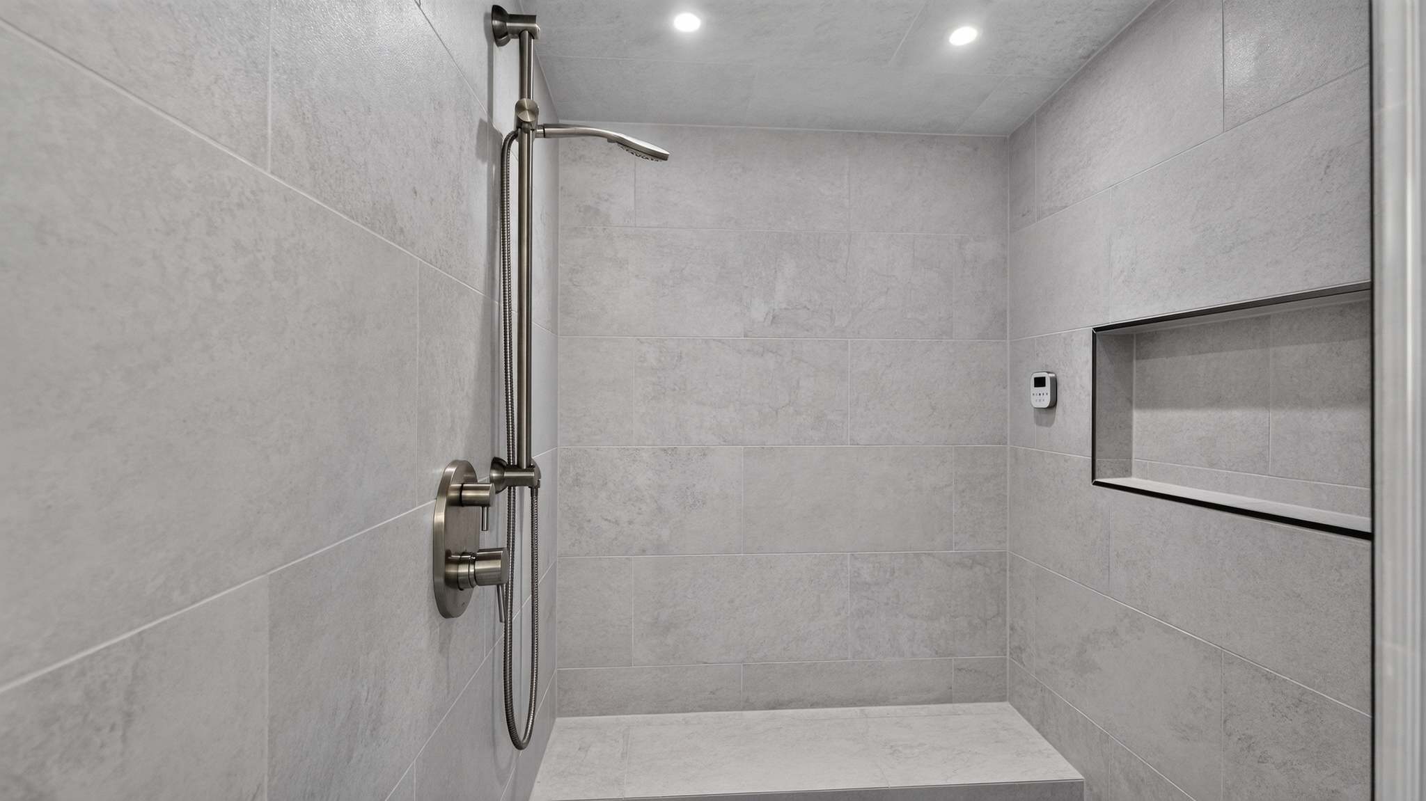 Close-up view of a modern walk-in shower featuring light gray large format tiles, a built-in bench, and a brushed nickel handheld shower on a slide bar.
