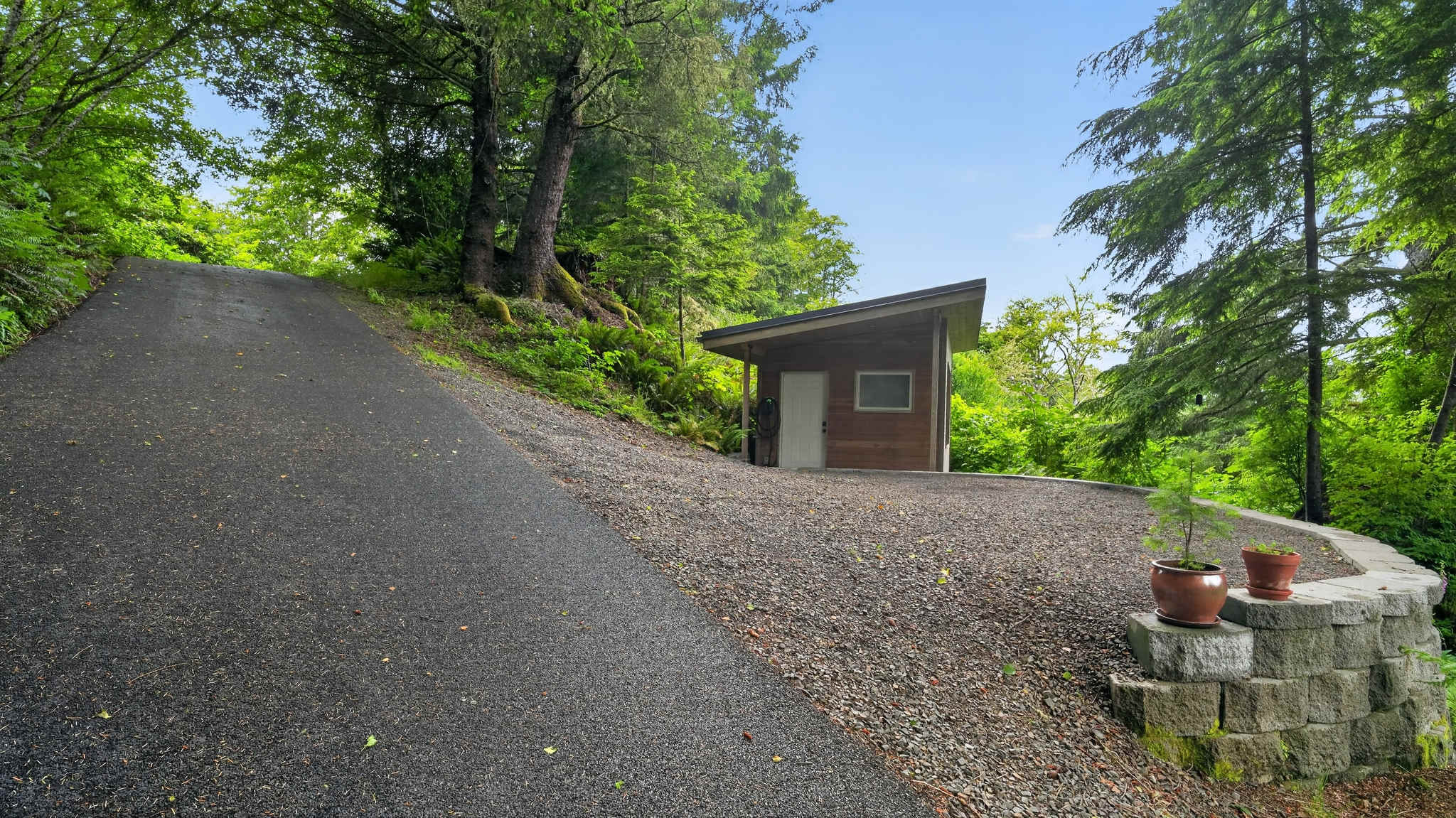 A steep asphalt driveway ascends to a gravel parking area, bordered by lush trees. A small, modern wooden outbuilding with a sloped roof and a white door stands on the right.