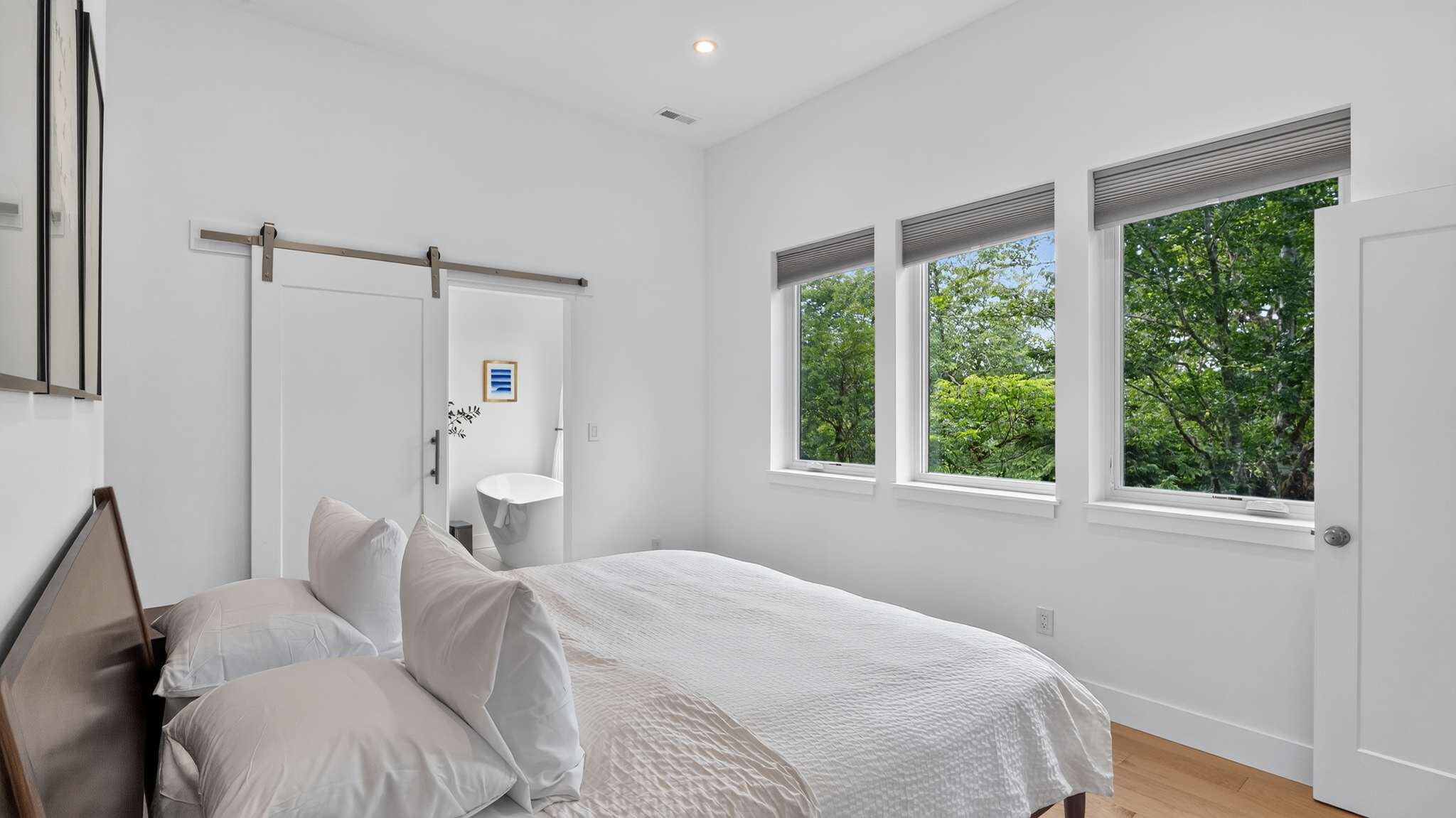 Modern bedroom with a white bed, light wood floors, and white walls, featuring three windows with tree views and a barn door leading to an en-suite bathroom.