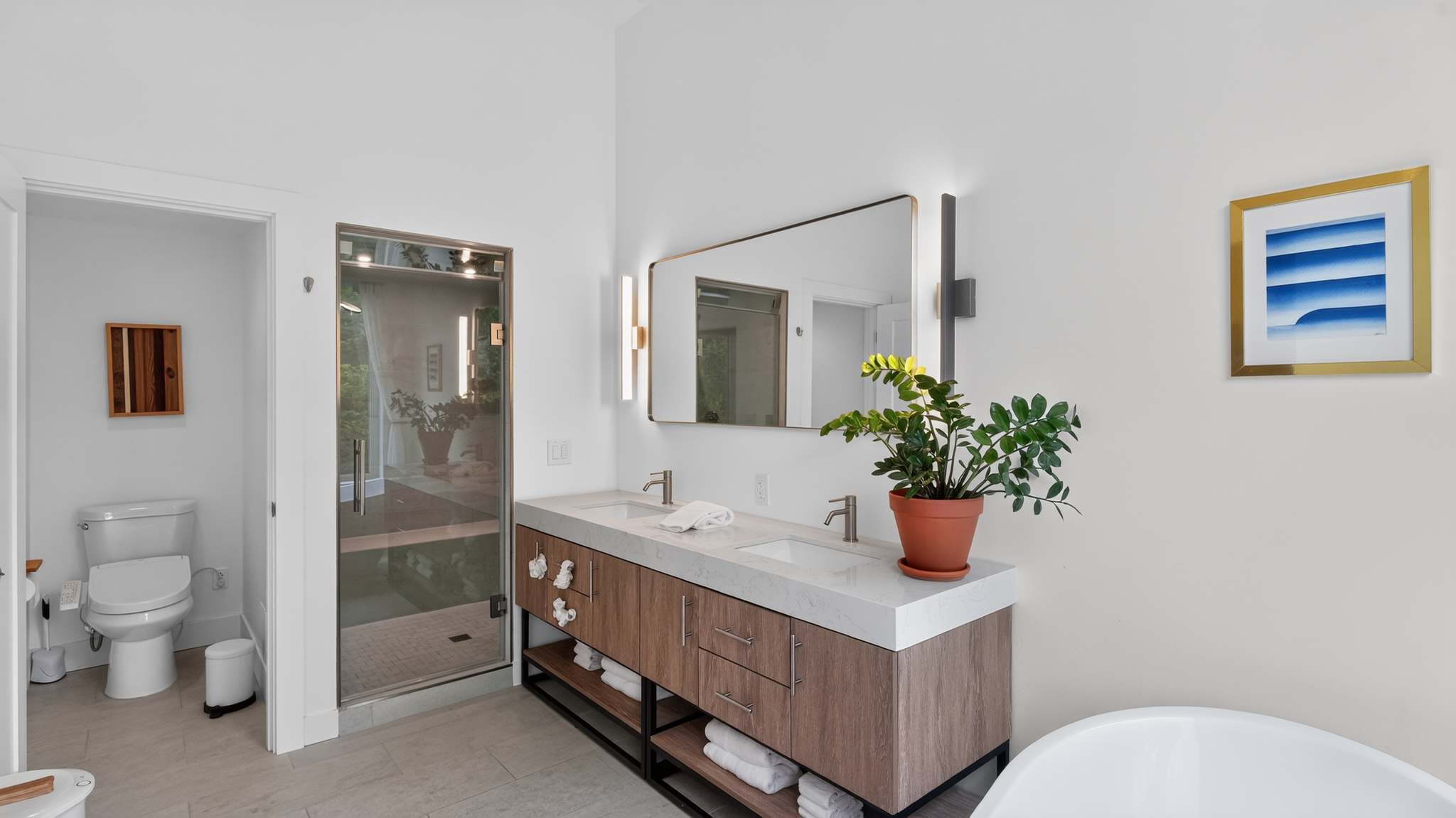 A spacious, modern bathroom features a double vanity with a light wood cabinet and white countertop, alongside a large freestanding white tub positioned by a window offering lush green views. A glass shower is visible to the left.