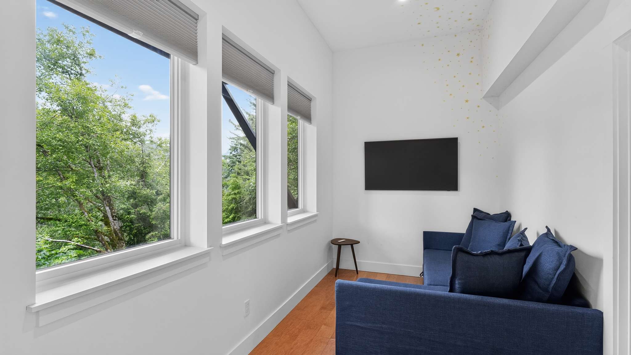 A cozy living space features a dark blue sofa with throw pillows, a wall-mounted TV, and three tall windows offering views of lush green trees. The room has light hardwood floors and white walls with subtle star decals.