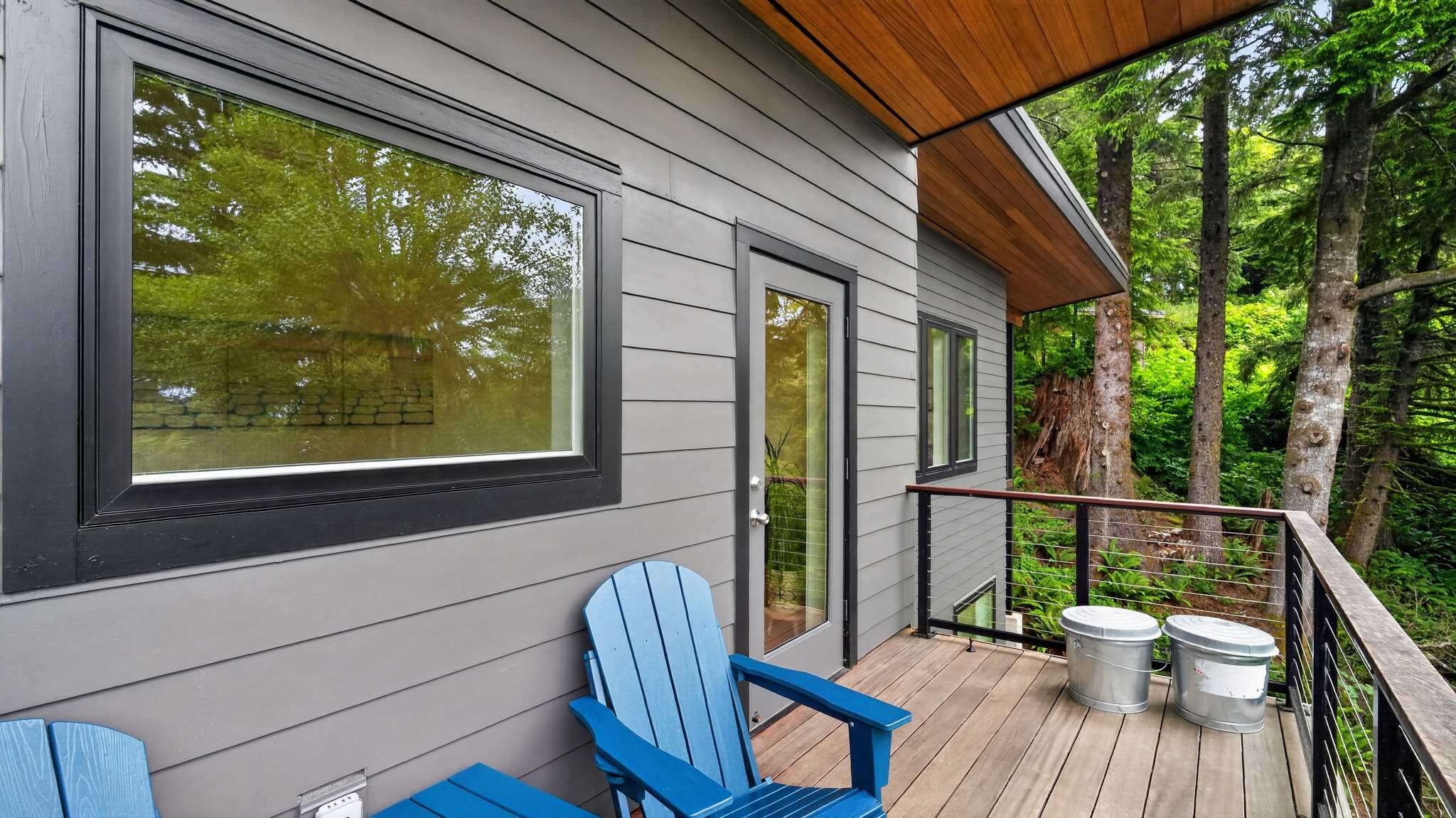 A cozy outdoor balcony features two bright blue Adirondack chairs and a small matching table, set on a wooden deck with black cable railings. The area is surrounded by lush green trees, offering a serene natural view.