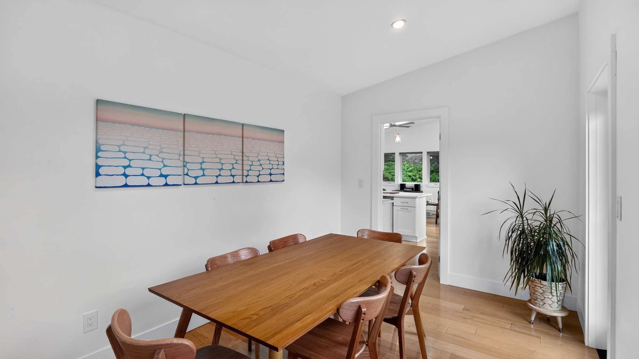 Contemporary dining room with a wooden table and chairs, light wood floors, and white walls, adorned with a large abstract artwork and offering a view into the adjacent kitchen.