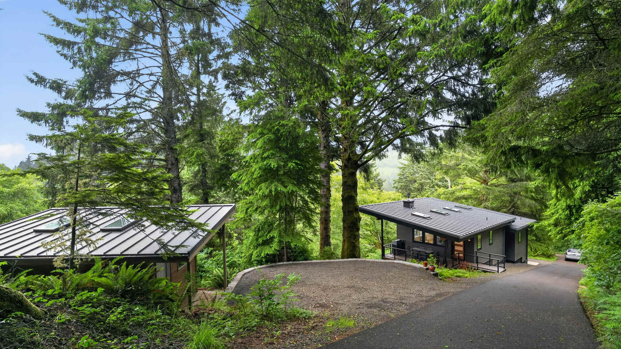 An aerial view reveals two modern, dark-roofed buildings nestled within a dense evergreen forest. An asphalt driveway leads to a gravel parking area between the main house and a smaller outbuilding.