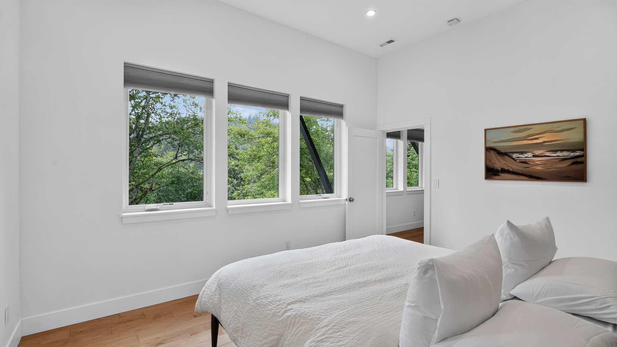 Bright bedroom with white walls, light wood floors, and a white bed, featuring three large windows offering lush green tree views. A framed artwork hangs on the wall.