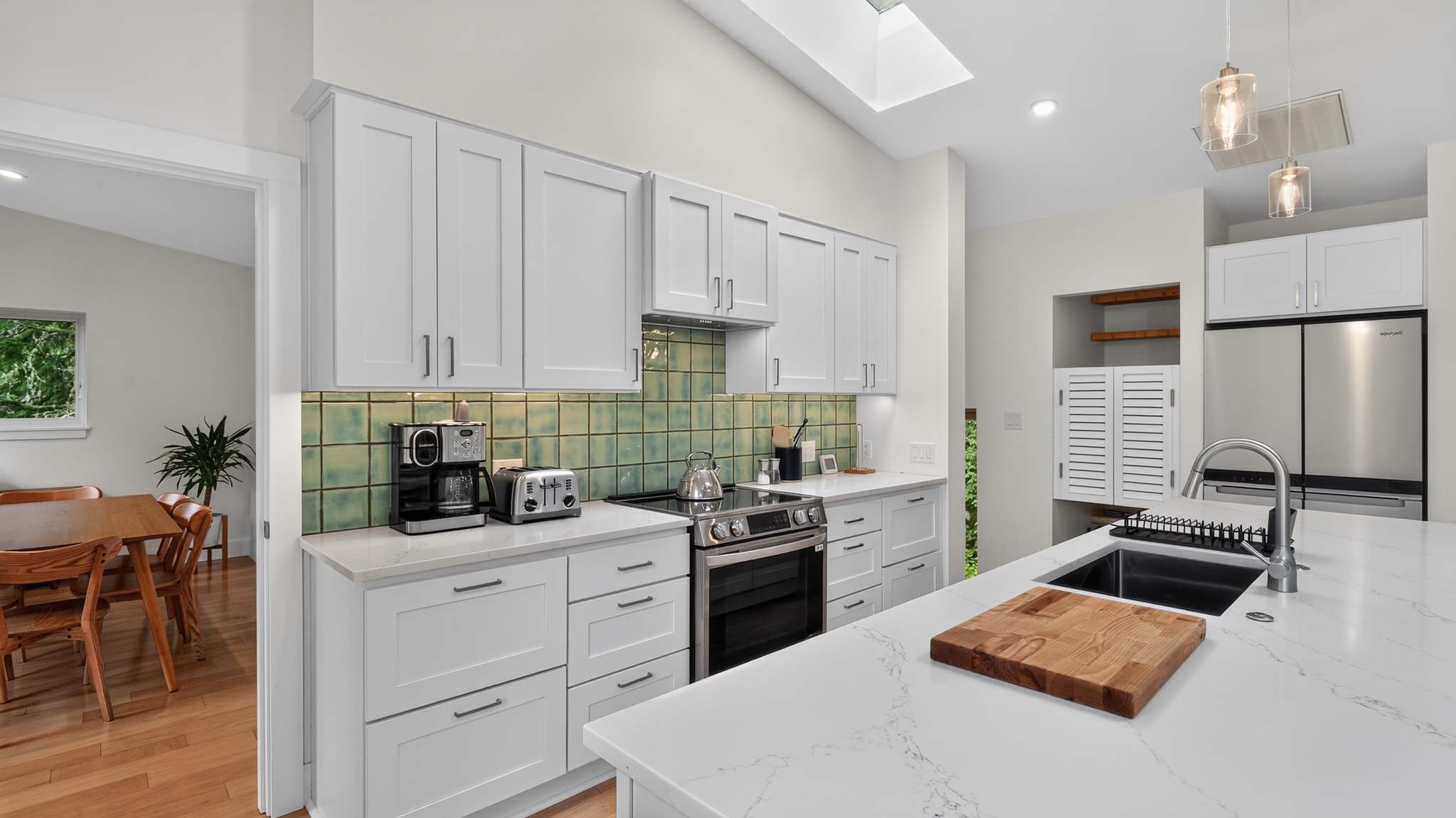 Modern kitchen with white shaker cabinets, white quartz countertops, a green tiled backsplash, and stainless steel appliances including a range and refrigerator. A large skylight brightens the space, and a doorway leads to a dining area.
