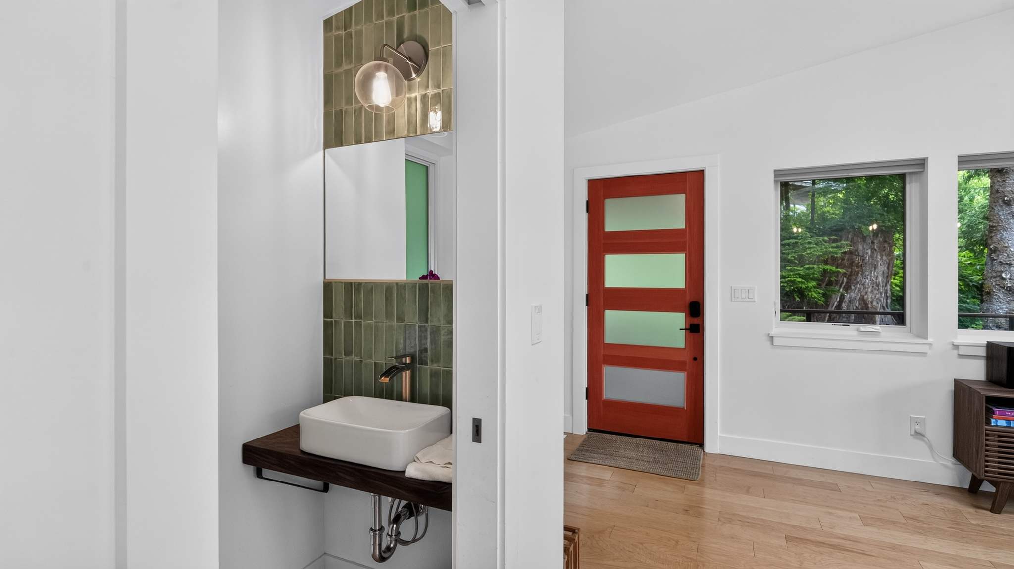 A modern half bathroom features a white vessel sink on a dark wooden floating vanity, with a gold faucet and a rectangular mirror above green subway tiles. A striking red front door with frosted glass panels is visible in the adjacent entryway.