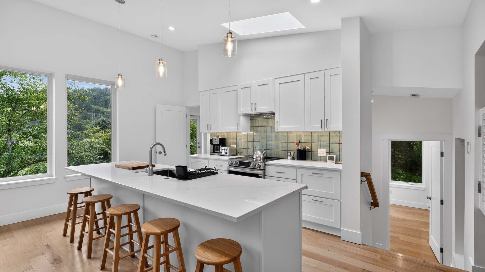 A modern kitchen with white cabinetry, light hardwood floors, and a large white quartz island with wooden bar stools. A skylight and large windows provide ample natural light and forest views, complementing the light green subway tile backsplash.