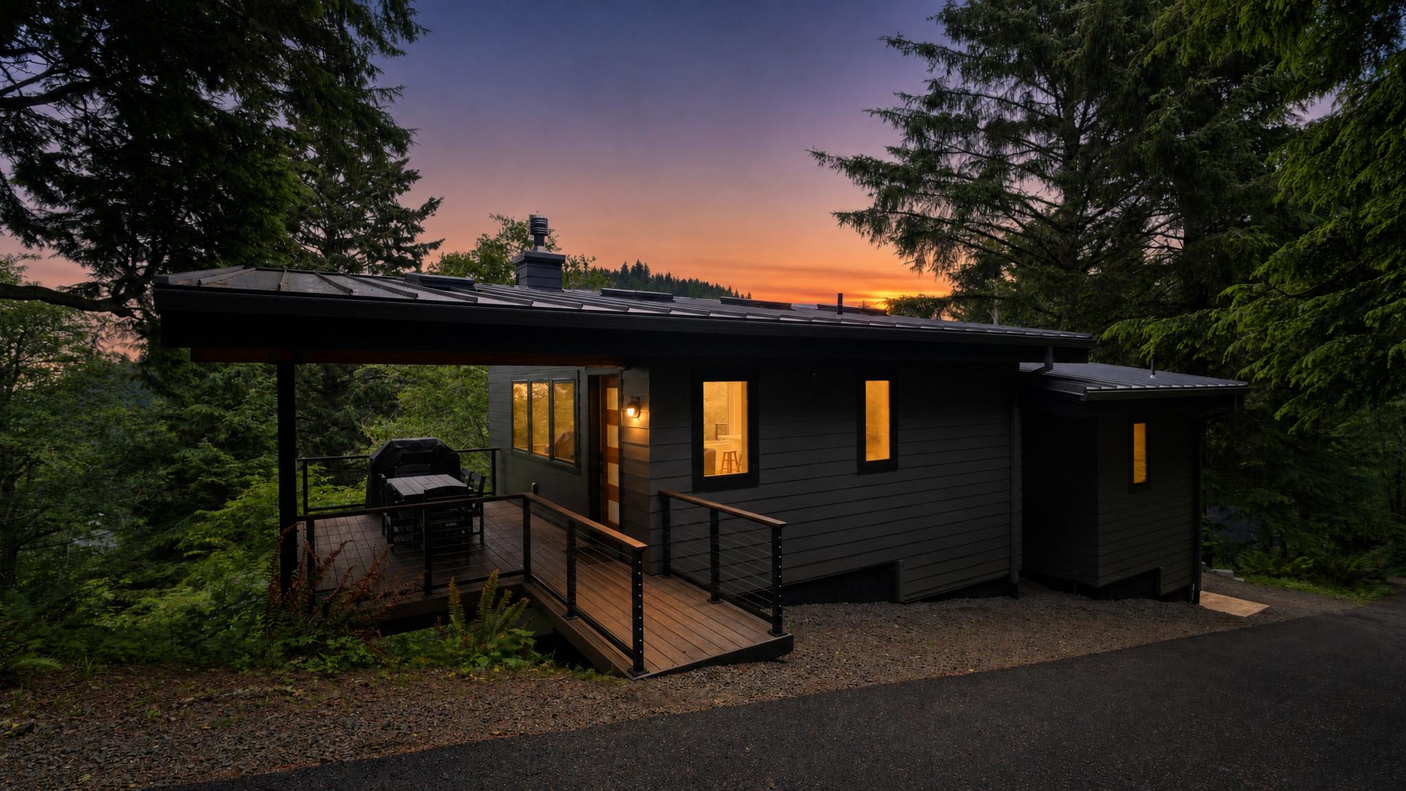 The front exterior of a modern dark gray house is illuminated at dusk, nestled among tall evergreen trees. A wooden ramp leads to a covered deck with a grill, creating an inviting entrance.