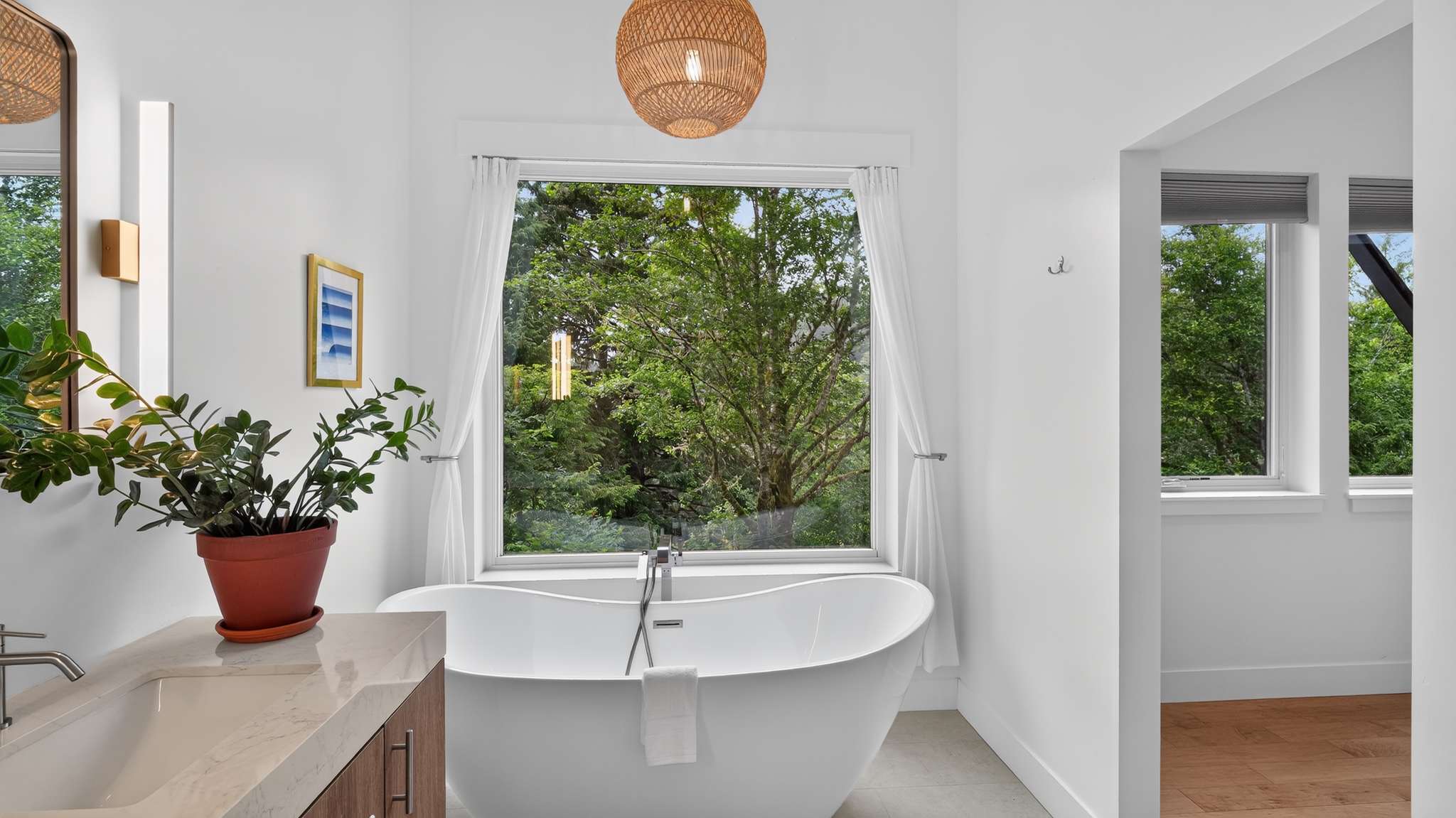 Bright bathroom featuring a freestanding white soaking tub positioned under a large window with lush green tree views, and a woven pendant light hanging from the ceiling.
