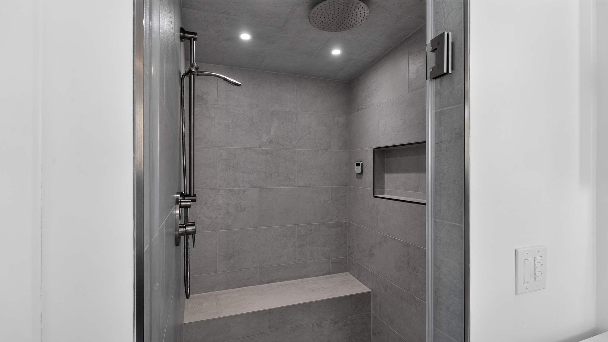 Modern walk-in shower with light gray large format tiles, a built-in bench, a shower niche, and both a rainfall showerhead and a handheld shower on a slide bar.