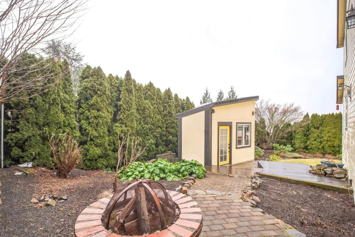 A backyard area featuring a circular brick fire pit with logs, surrounded by a mix of grass and garden beds, with a small yellow shed and tall evergreen trees in the background.