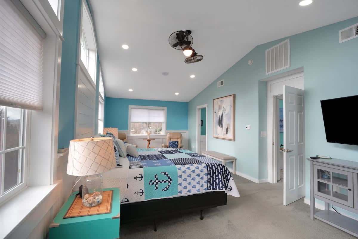 Another perspective of the light blue bedroom, featuring a gray media console with a flat-screen TV, the bed with its blue quilt, and a doorway leading to another room. The sloped ceiling and recessed lights are prominent.