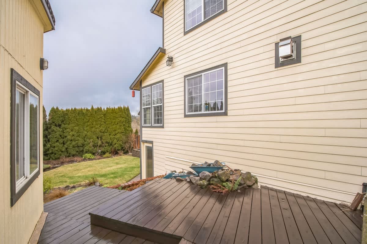The back of a light yellow house with multiple windows and a dark wooden deck. A small rock garden feature is visible on the deck, overlooking a grassy yard.
