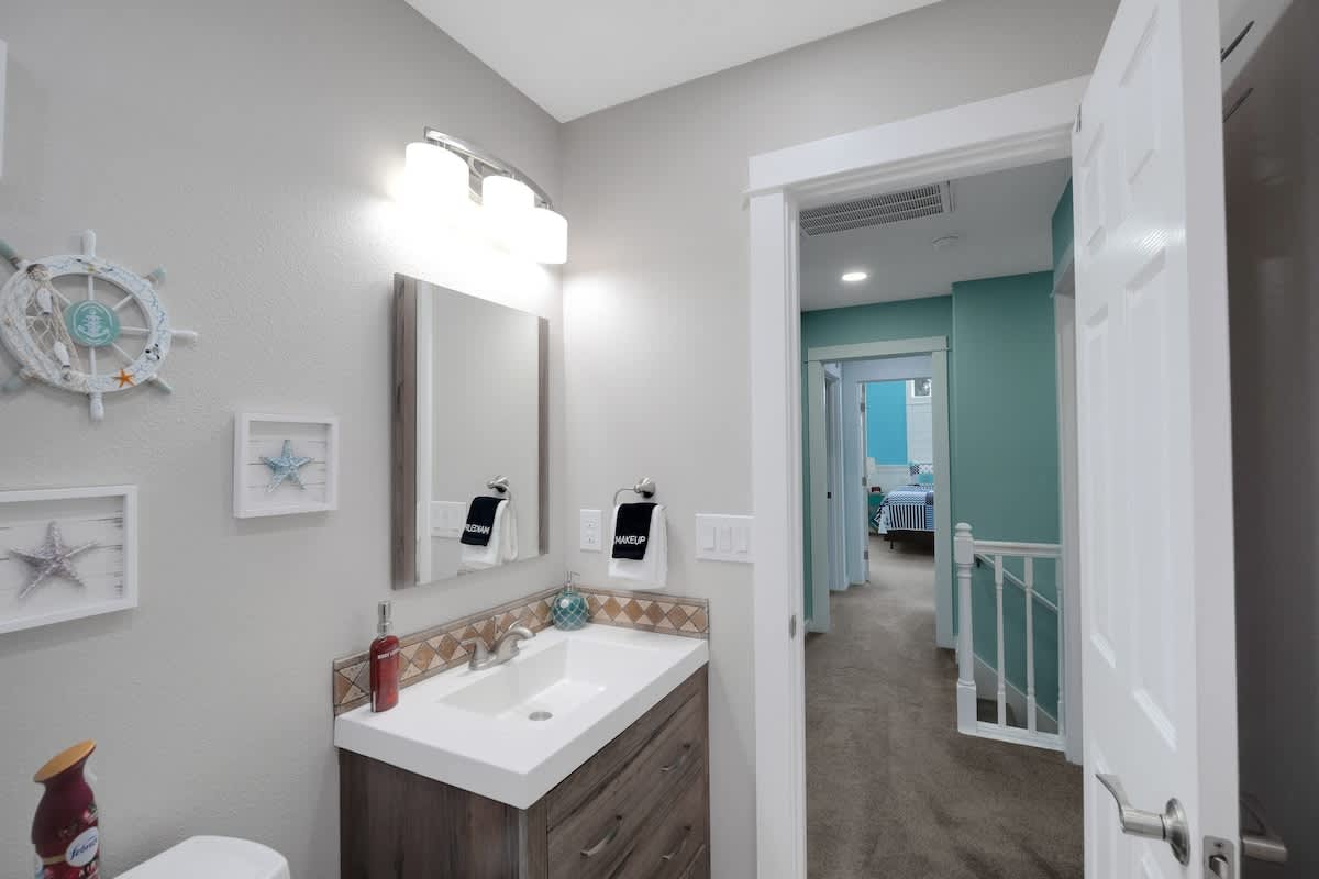 A modern bathroom features a dark wood vanity with a white sink, a rectangular mirror, and a three-bulb light fixture, complemented by light gray walls and coastal-themed decor. A hallway leads to other rooms.