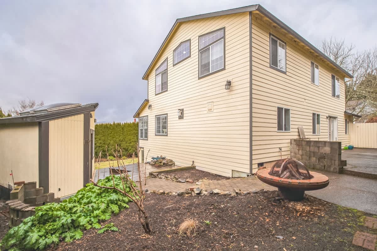 A wide view of the back of a light yellow house with a dark trim, featuring a paved area, a fire pit, and a small shed. A lush green hedge borders the property.