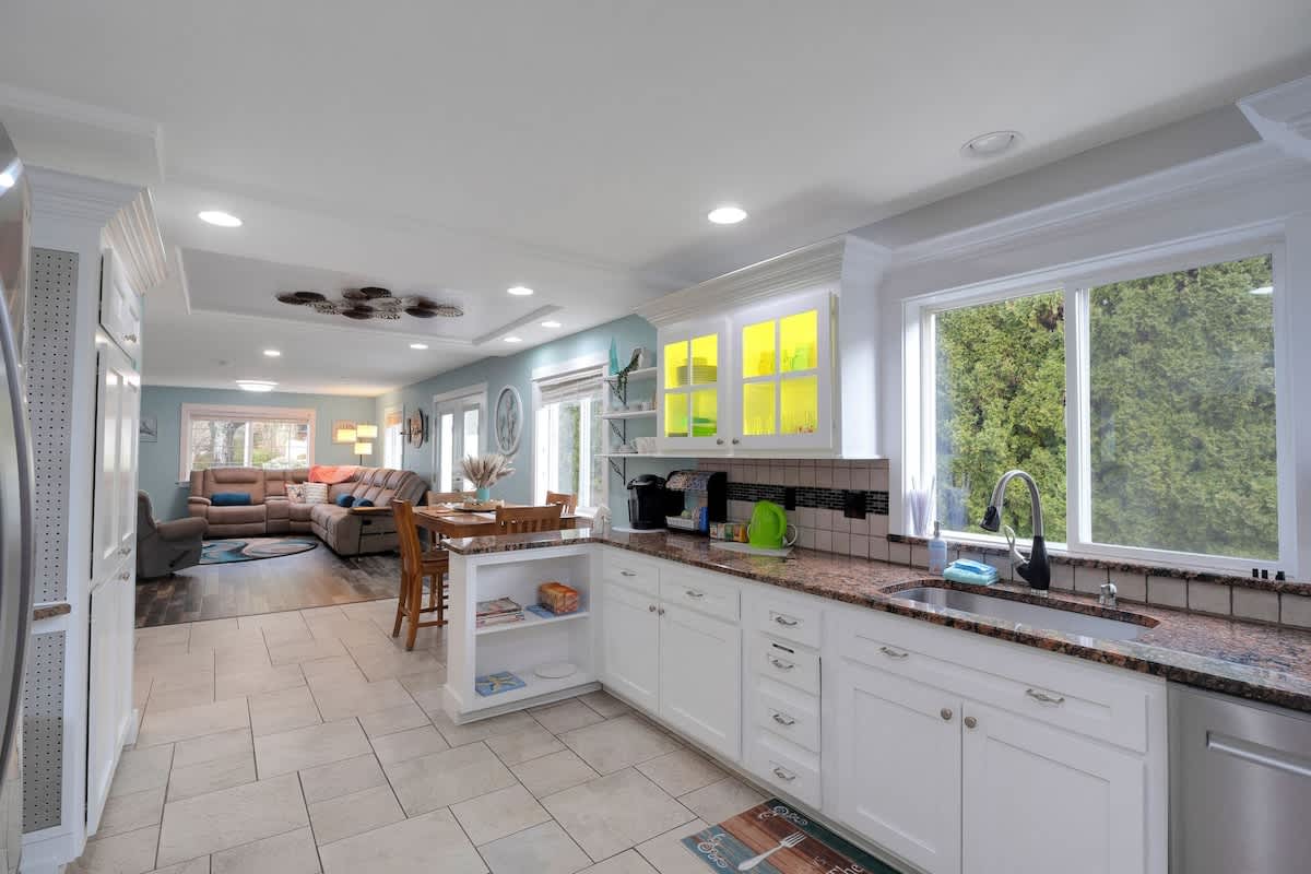 Open-concept kitchen with white cabinets, granite countertops, and light tile flooring, featuring a coffee station with a Keurig machine and an open view to the adjacent dining and living areas.
