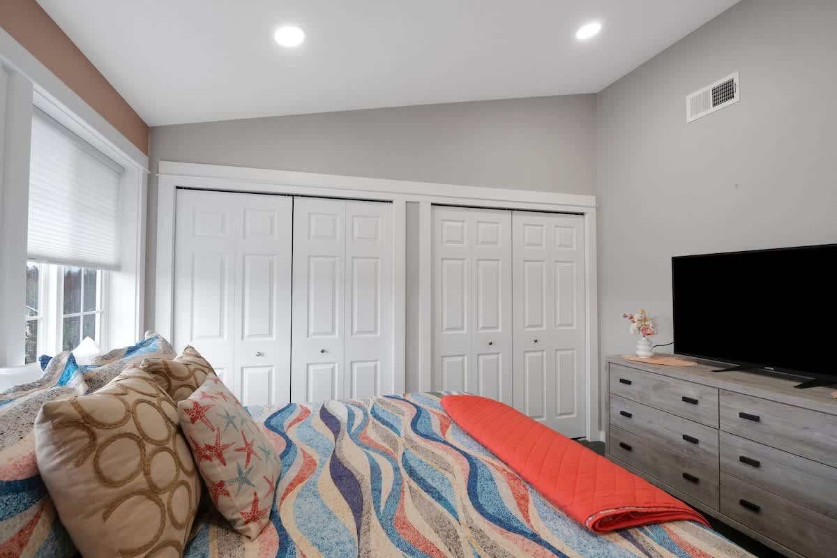 This angle of the bedroom highlights the bed with its colorful quilt, the windows with white blinds, and a wall of white bi-fold closet doors. The room has a sloped ceiling and recessed lighting.
