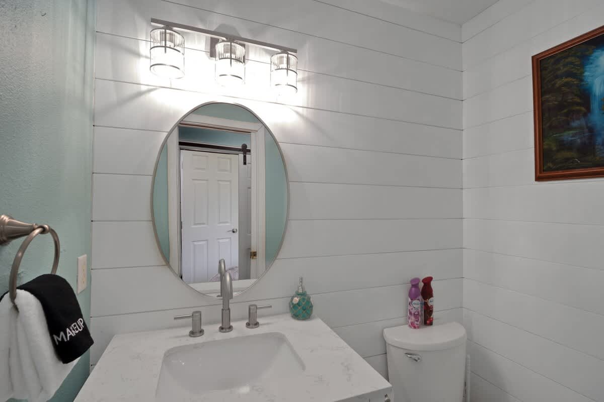 A compact bathroom with a white shiplap accent wall, a round mirror above a single vanity with a white countertop, and a toilet visible to the right.