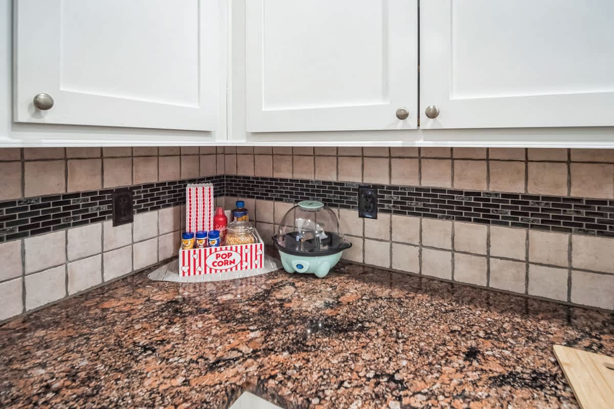 A close-up of a kitchen corner with granite countertops, white upper cabinets, and a tiled backsplash. A popcorn machine and popcorn box are visible on the counter.
