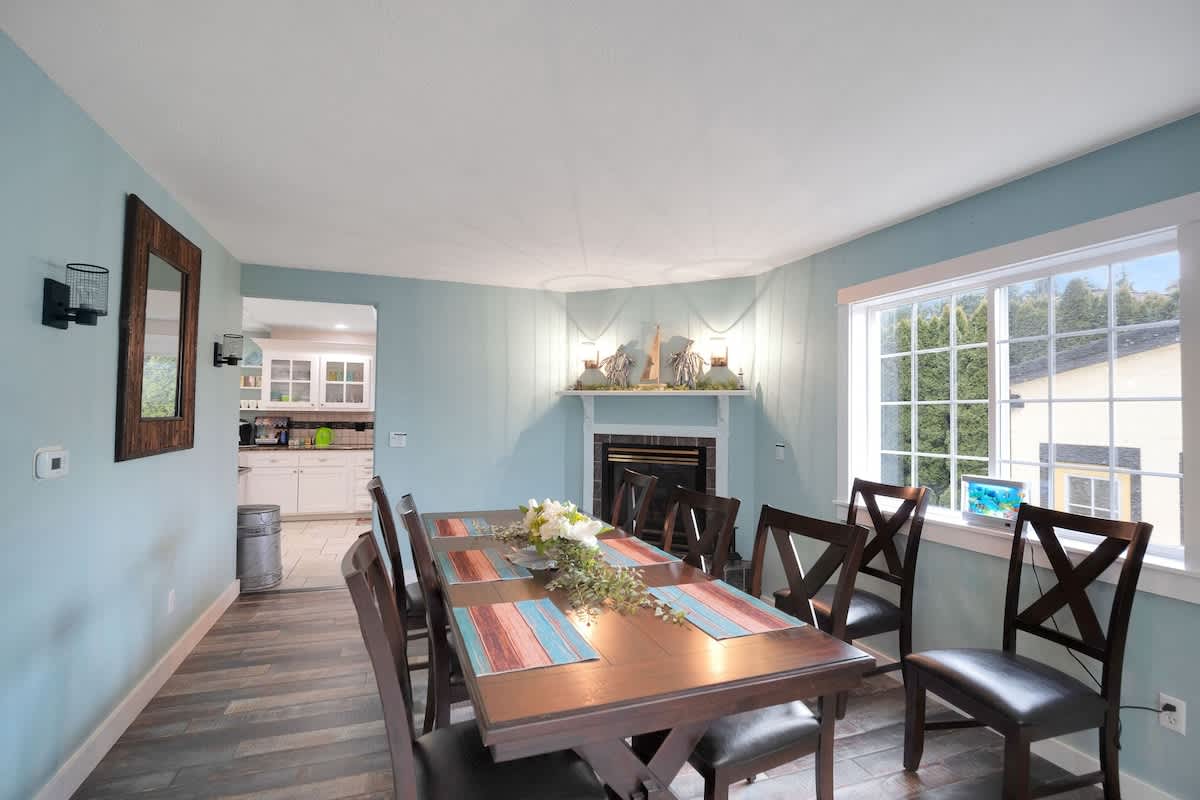 A spacious dining room with light blue walls, a dark wood dining table, and matching chairs. A corner fireplace with a white mantel and decorative items is visible in the background.