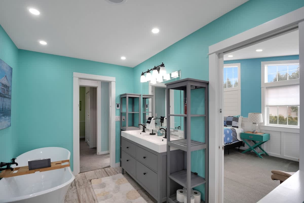 The double vanity area of the teal-walled bathroom, featuring a gray cabinet with two sinks, white countertops, black faucets, and an open shelving unit for storage.