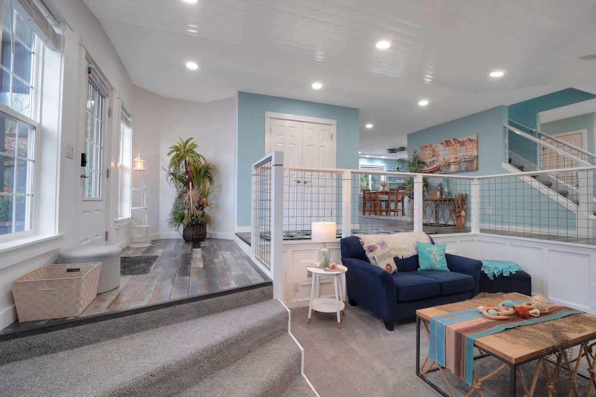 A split-level living room features a navy blue sofa on gray carpet, separated by a white railing with wire mesh from an upper landing with wood-look flooring and an entrance. Light blue walls and recessed lighting complete the bright space.