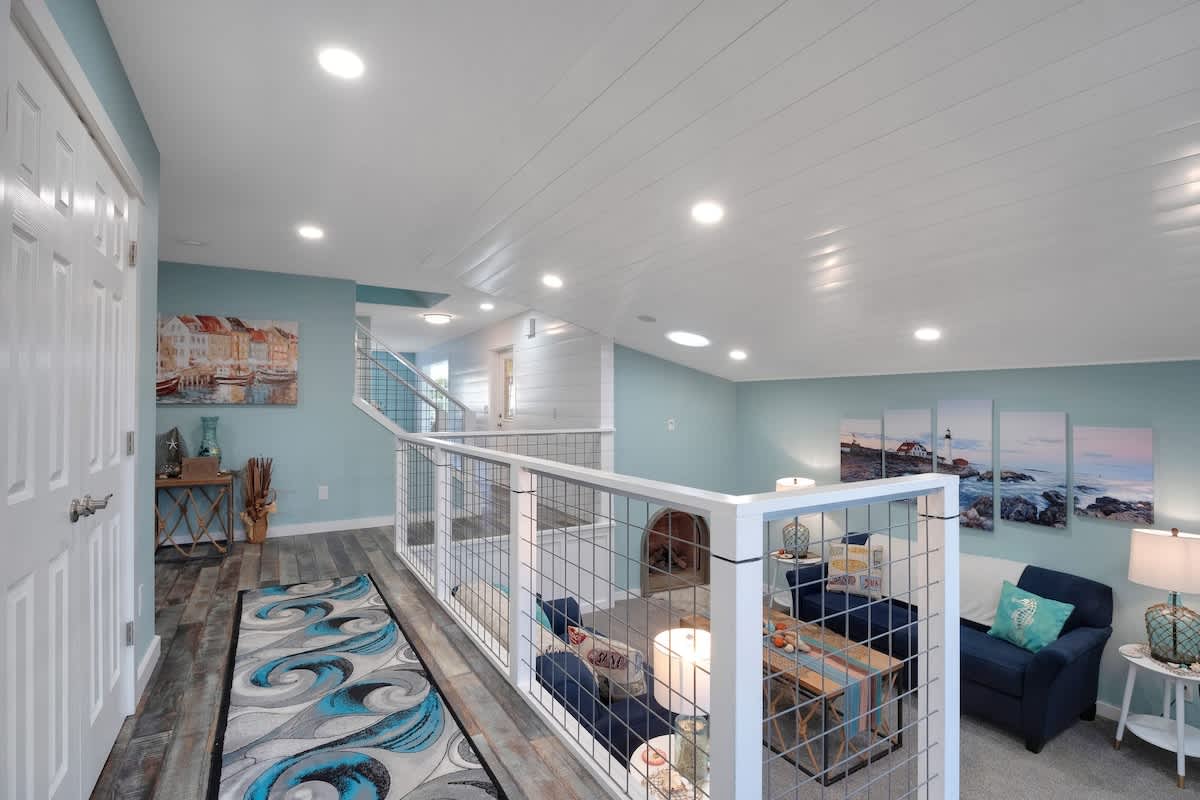 Upper landing area with light blue walls and wood-look flooring, featuring a modern metal and wood railing overlooking a lower living space with a sofa and coastal artwork.