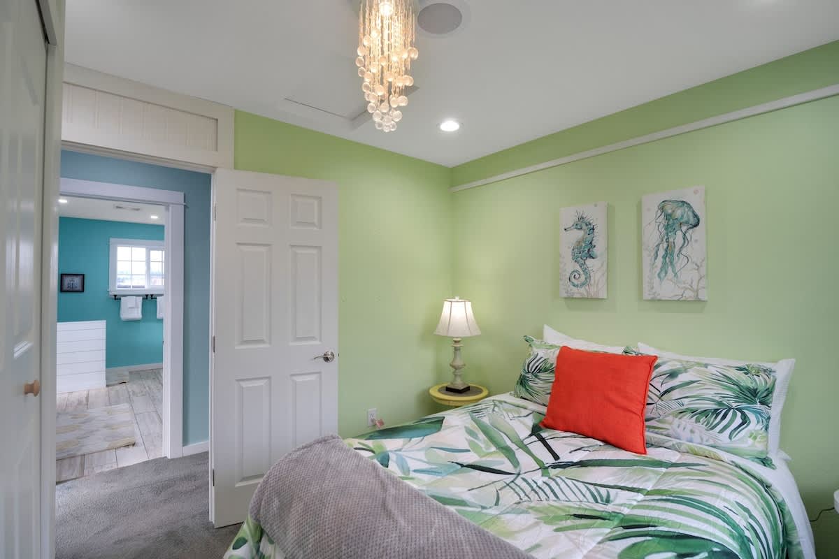 This view of the light green bedroom shows the bed with tropical bedding, a nightstand with a lamp, and a doorway leading to a light blue bathroom. Nautical wall art and a shell chandelier are visible.