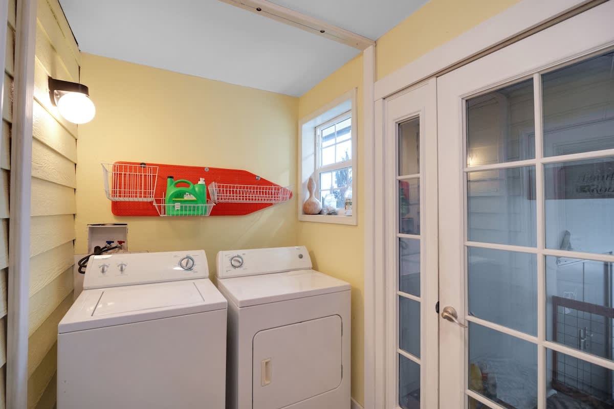 A compact laundry room features bright yellow walls, a white washer and dryer set, and a wall-mounted red ironing board with a storage basket, illuminated by a window and an overhead light.