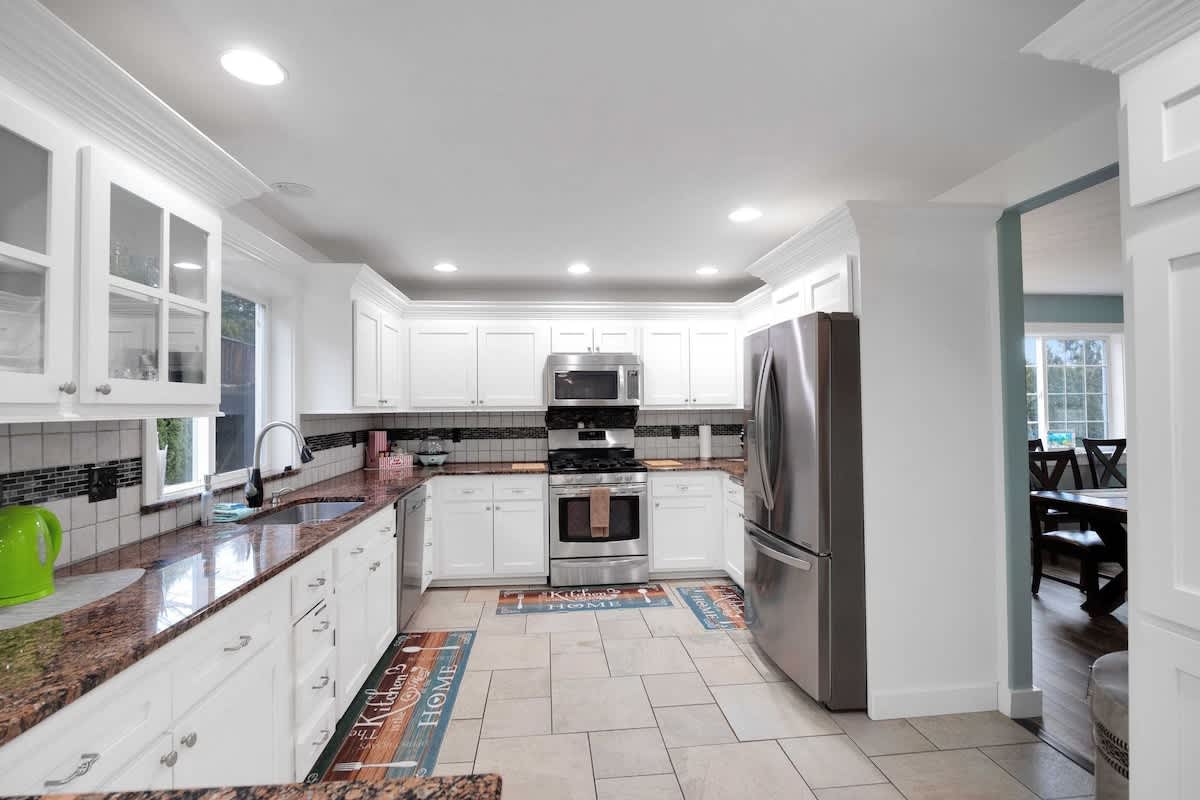 A spacious kitchen with white shaker-style cabinets, granite countertops, and stainless steel appliances including a refrigerator, oven, and microwave. Light tile flooring completes the look.