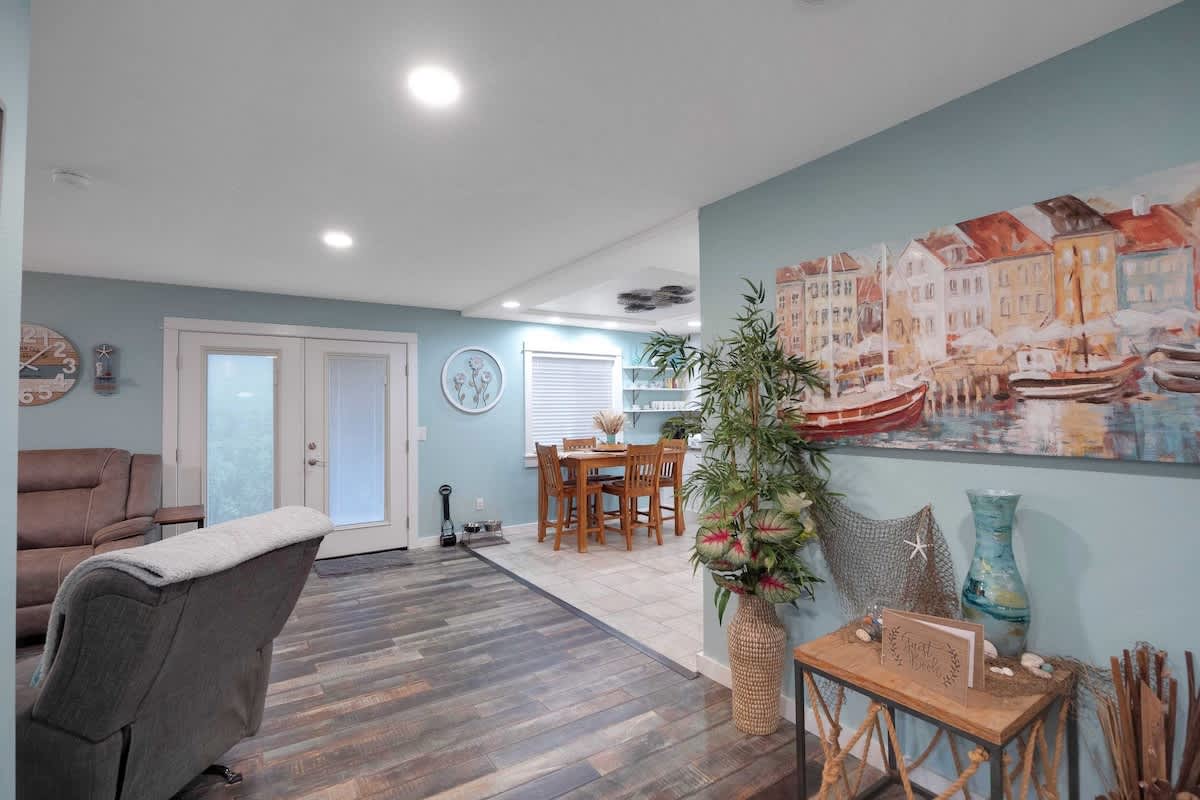 A transitional space with light blue walls and dark wood-look flooring, featuring a large nautical-themed painting, a decorative plant, and an open view towards a dining area and kitchen.
