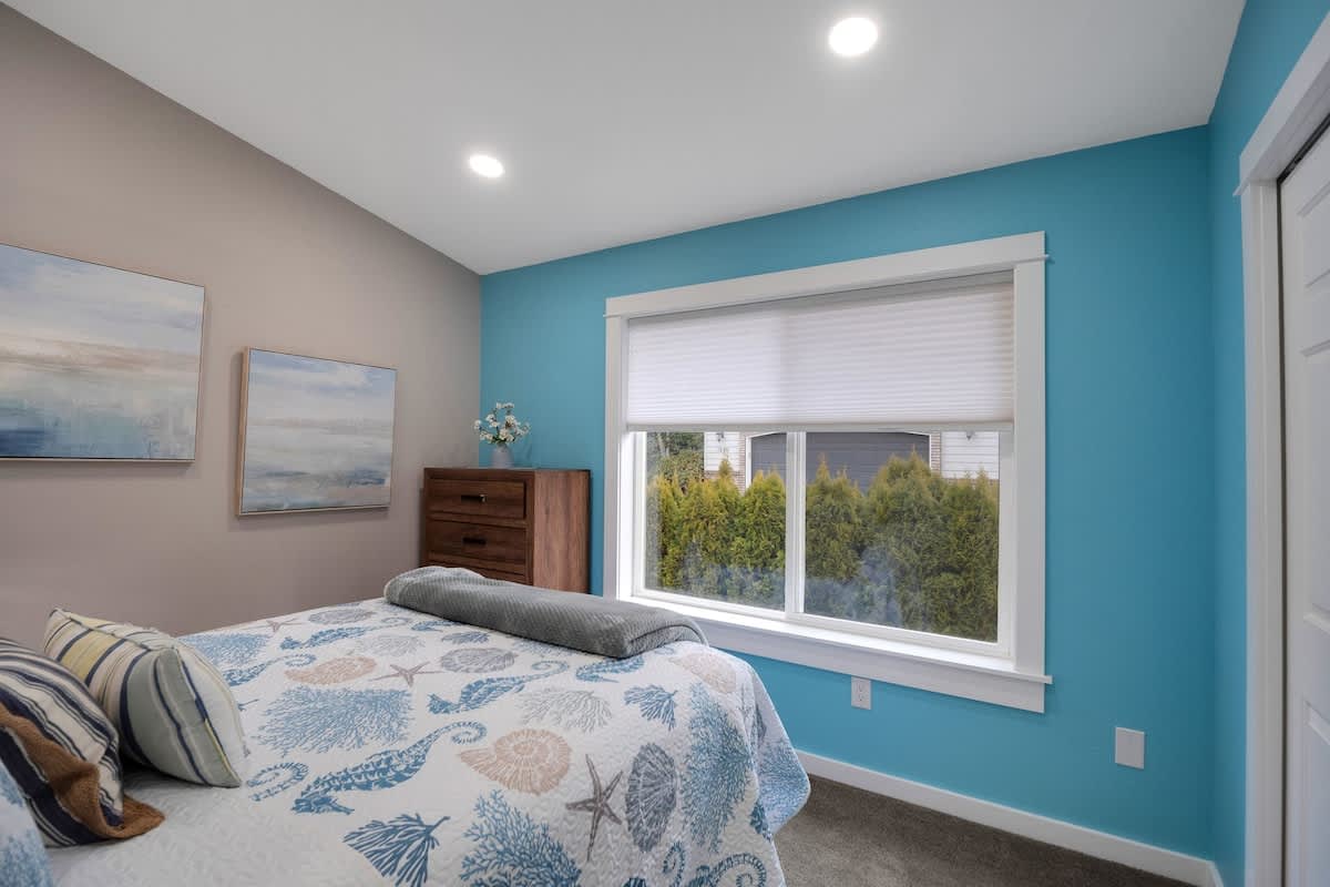A cozy bedroom featuring a blue accent wall and a gray wall, a queen-sized bed with blue and white ocean-themed bedding, and a large window with white blinds.