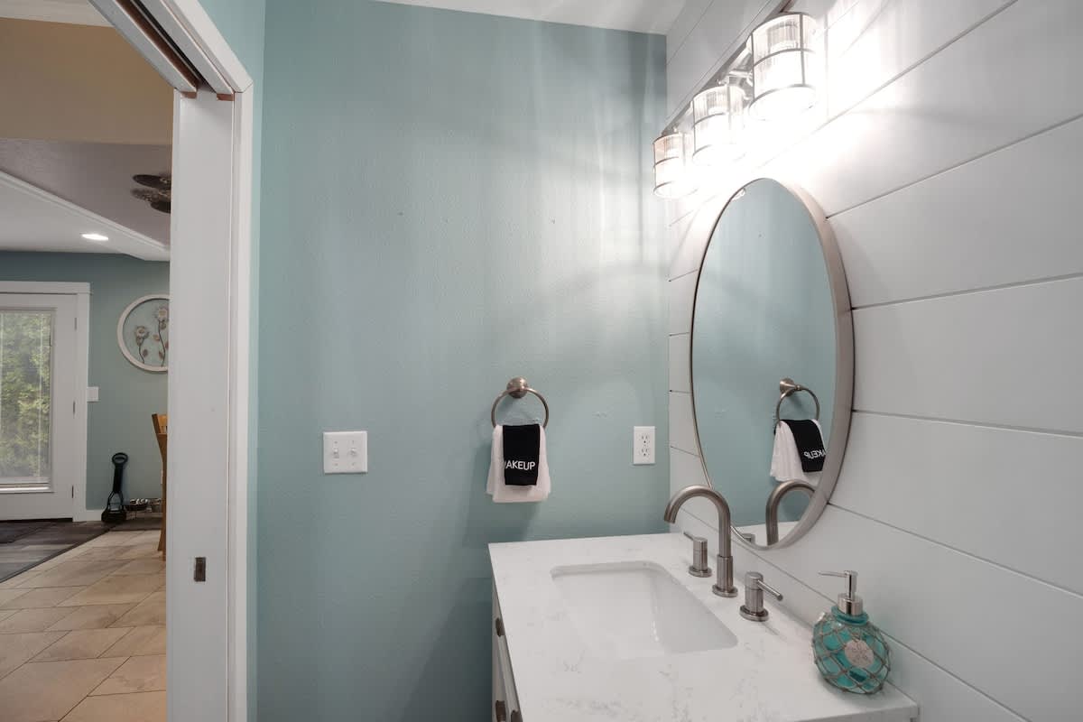 A wider view of the compact bathroom, showcasing the white shiplap wall, round mirror, single vanity, and the light blue painted wall contrasting with the shiplap.