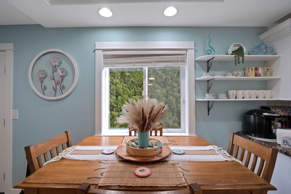 A dining area with light blue walls, a wooden dining table set with a runner and centerpiece, and a window overlooking greenery, flanked by white floating shelves with dishware.