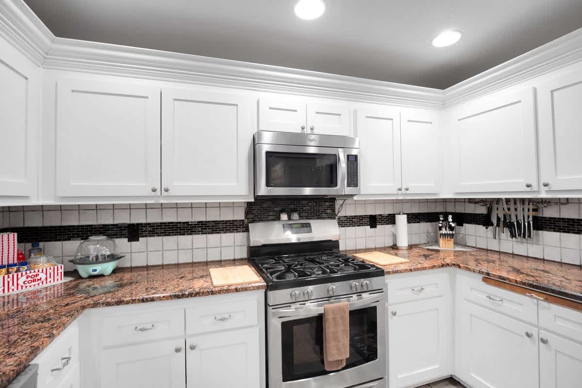 A close-up of the kitchen's cooking area, featuring a stainless steel gas range with an overhead microwave, white cabinets, and a tiled backsplash with a decorative black border.