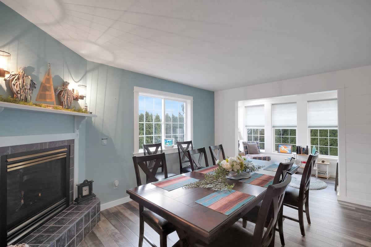 A spacious dining room featuring a large dark wood dining table with seating for eight, a tiled fireplace, and multiple windows providing natural light. The walls are painted a light blue with white wainscoting.