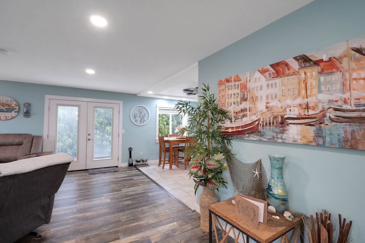 A bright living area features light blue walls, dark wood-look laminate flooring, and a large coastal-themed painting, with a dining nook visible through an archway and French doors leading to the outside.