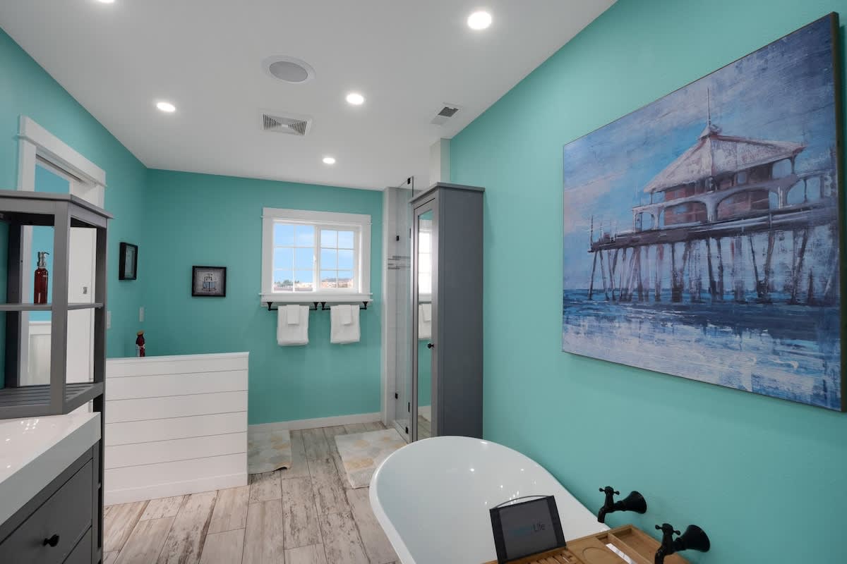 A wide view of the spacious bathroom with teal walls, a freestanding tub, light wood-look floor tiles, and a white shiplap-style half-wall with a window above it.