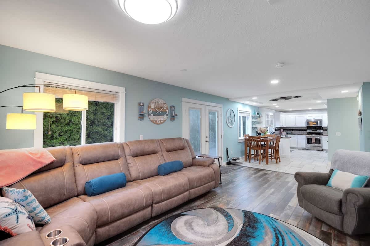 Spacious living room with a large brown sectional sofa, dark wood-look flooring, and light blue walls, featuring multiple windows with blinds and an open view to the kitchen and dining area.
